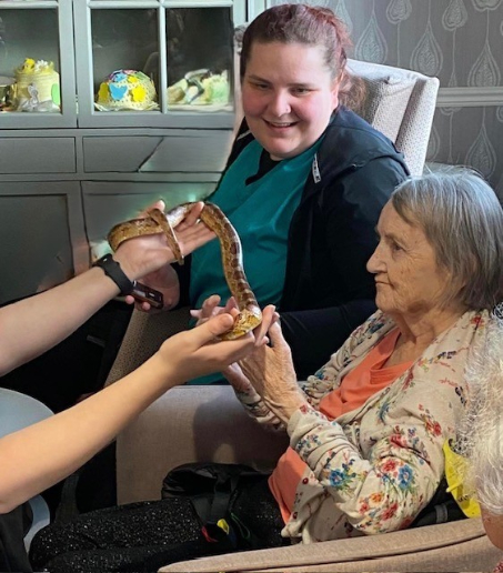 Kit, a resident at Haddington Care Home, smiling as she holds a snake during her 80th birthday celebration with ZooLabs, surrounded by staff, residents, and loved ones