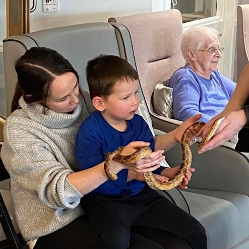 Kit, a resident at Haddington Care Home, smiling as she holds a snake during her 80th birthday celebration with ZooLabs, surrounded by staff, residents, and loved ones