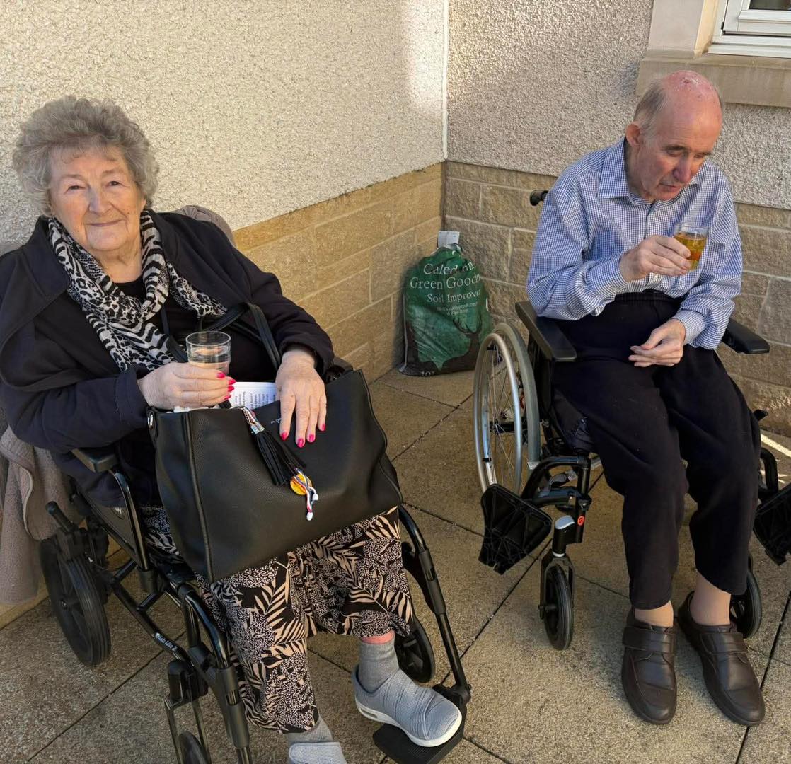 older lady and an older man sat in wheelchairs outside with drinks